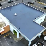 Overhead view of a flat commercial building roof with a fresh blue coating, featuring ventilation fixtures, surrounded by a parking area with cars, in an urban environment.