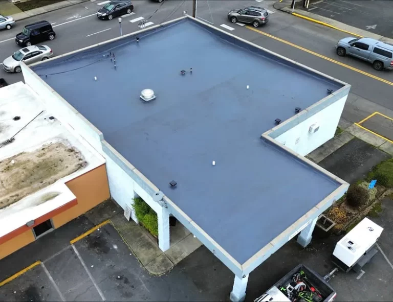 Overhead view of a flat commercial building roof with a fresh blue coating, featuring ventilation fixtures, surrounded by a parking area with cars, in an urban environment.