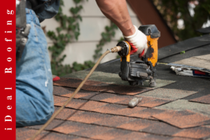 A worker kneels on a rooftop using a nail gun to secure shingles, wearing jeans and gloves. A red vertical banner reads "iDeal Roofing."
