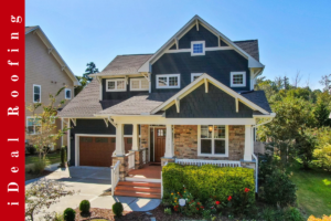 Charming two-story house with dark siding and stone accents. Features a well-kept yard and vibrant flowers. Bright, sunny day with clear skies. "iDeal Roofing" text vertically on a red banner to the left.