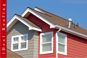 Modern house with red and gray siding and a brown gabled roof under a clear blue sky, showcasing homes needing roof repair vs roof replacement, with "iDeal Roofing" displayed vertically on the left.