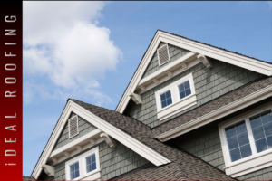 Residential house roof with gray asphalt shingles, gable design, and white-trimmed windows under a blue sky, highlighting roofing structure and exterior home details.