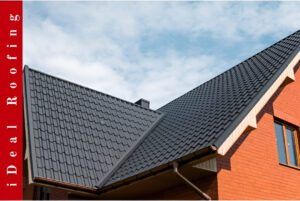 Brick house with black tiled roofs under a clear blue sky, promoting roof repair vs roof replacement services, with vertical text reading "iDeal Roofing" on a red background.