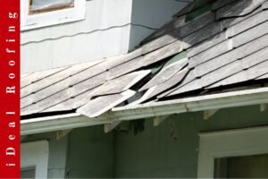 Old, weathered shingles are visible on a house roof, with some lifted and damaged, showing conditions that often lead to emergency roof repair needs. The text “iDeal Roofing” appears on the left in red.