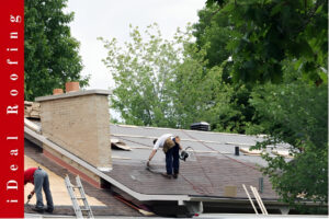 Two workers perform emergency roof repair on a house roof surrounded by green trees, with one using a nail gun while the other prepares materials. A brick chimney is visible in the calm setting.