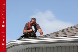 A roofer wearing safety gear performs emergency roof repair using a nail gun on a shingled roof under a clear sky, with a vertical banner reading “iDeal Roofing” in red.