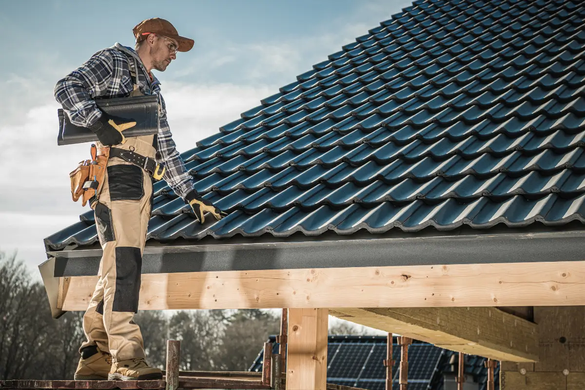 A roofer in work clothes and a cap inspects a dark gray tile roof, holding a clipboard and wearing a tool belt