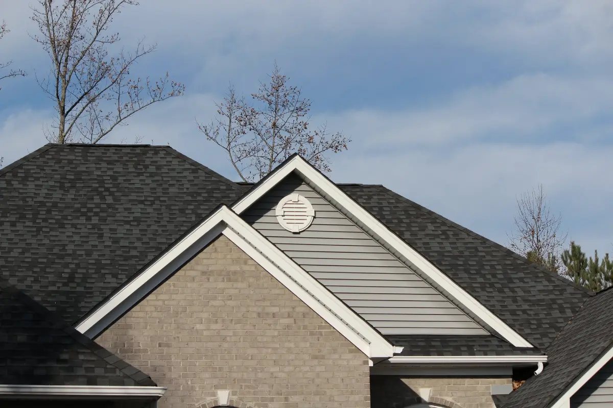 A section of a house featuring dark gray asphalt shingles on the roof, light gray siding in the gable, and brickwork below. Bare tree branches are visible against a cloudy blue sky with scattered clouds