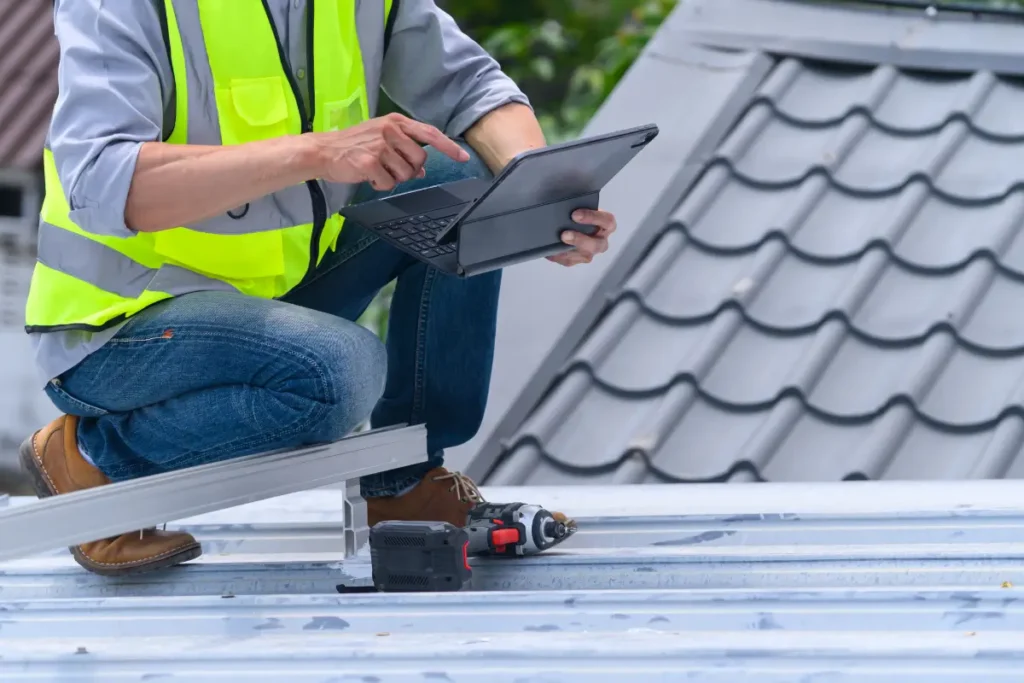 Roofing contractor crouching on a metal roof while using a digital tablet.