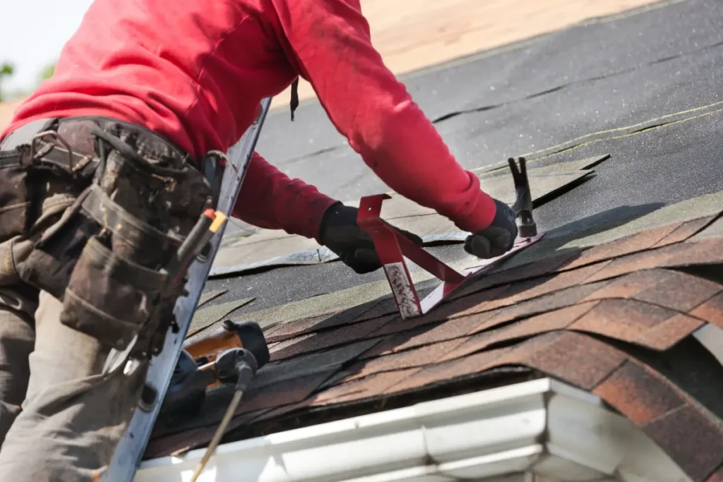 A person wearing a red long-sleeved shirt and tool belt hammers shingles onto a roof.