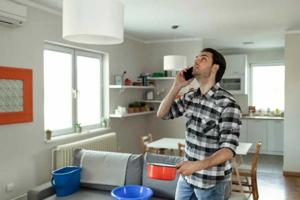 Concerned homeowner holding a pot and talking on the phone while catching water leaking from the ceiling.