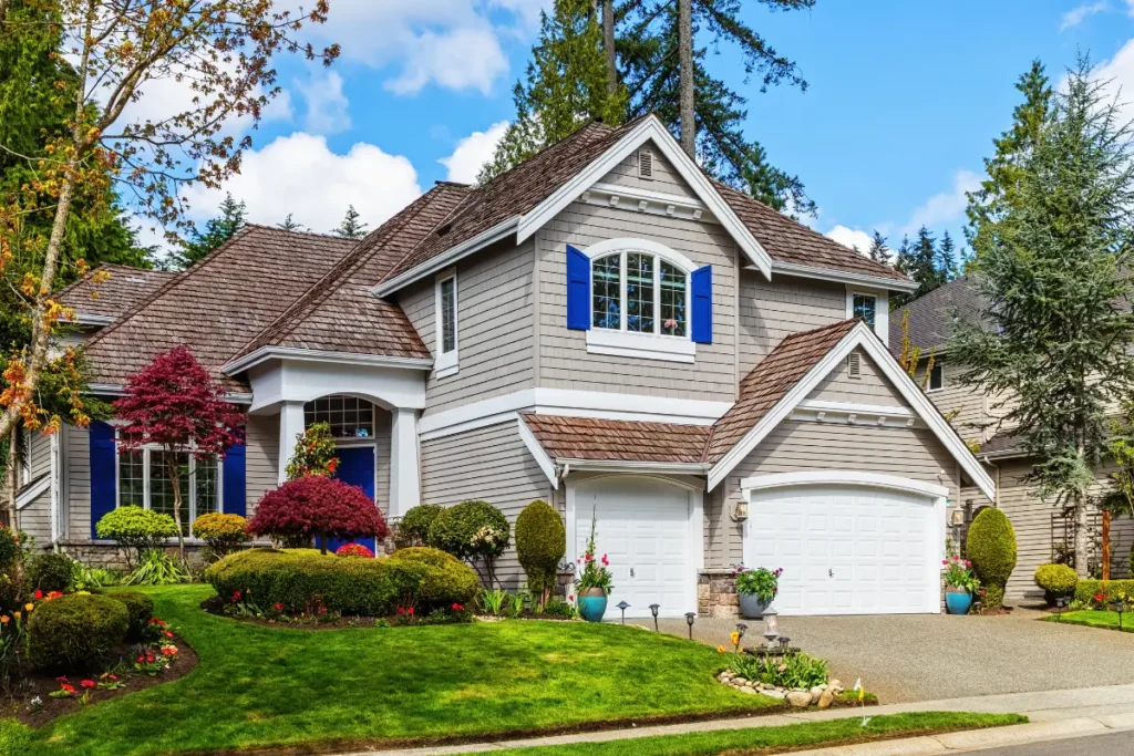 A large, two-story gray house with a brown roof, blue shutters, a well-manicured lawn, and colorful landscaping.