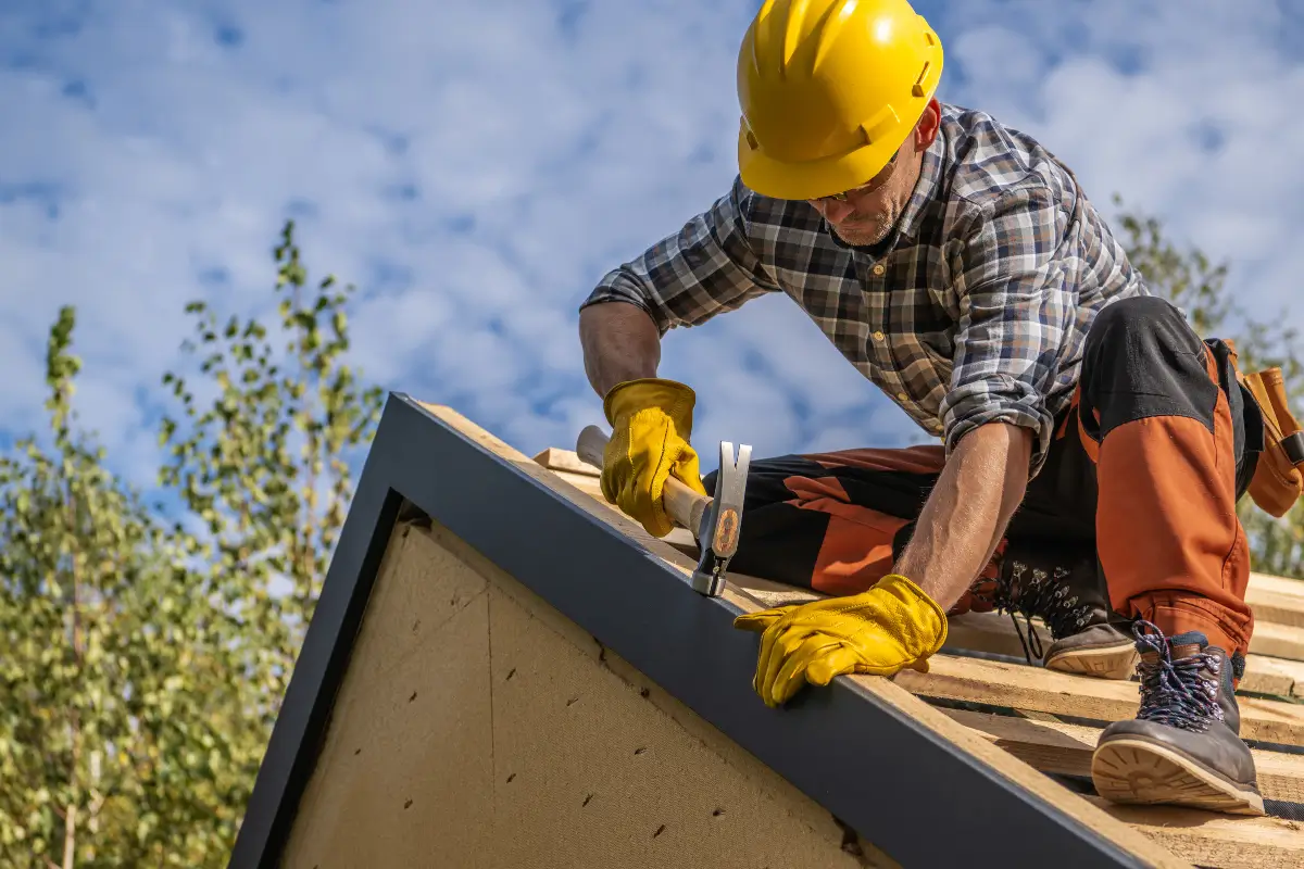 Professional roofer wearing a yellow hard hat and safety gloves hammering a metal edge flashing onto a residential roof structure under a blue sky