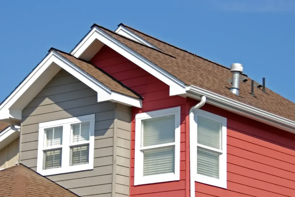 Exterior roofline of a residential house featuring brown asphalt shingles, red and grey siding, and white window trim.