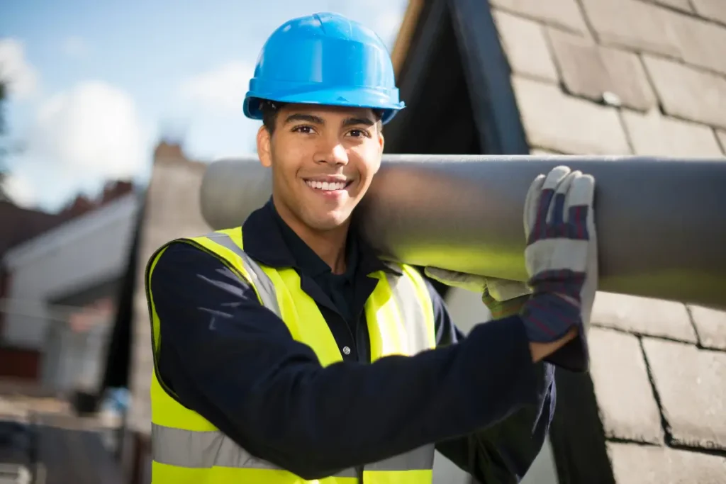 Smiling male roofer wearing a blue hard hat and high-visibility safety vest carrying roofing felt materials.