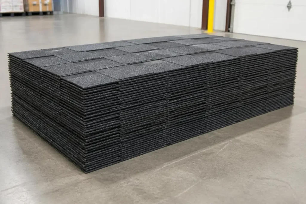 A large stack of dark gray or black asphalt roofing shingles neatly bundled on a warehouse factory floor, prepared for shipment or covered by a standard manufacturer warranty.