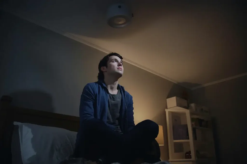 An anxious young man sitting cross-legged on a bed at night, staring up at a ceiling light fixture with a worried expression, illustrating the stress of household property issues.