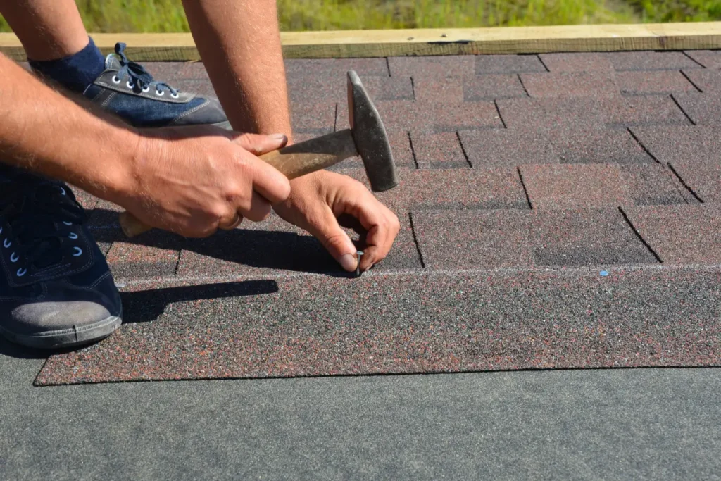 Roofer using a hammer to secure asphalt shingles onto a roof during installation.