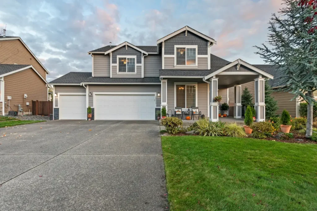Two-story craftsman home featuring grey siding, dark asphalt shingles, white garage doors, and a covered front porch with a manicured lawn under a twilight sky.