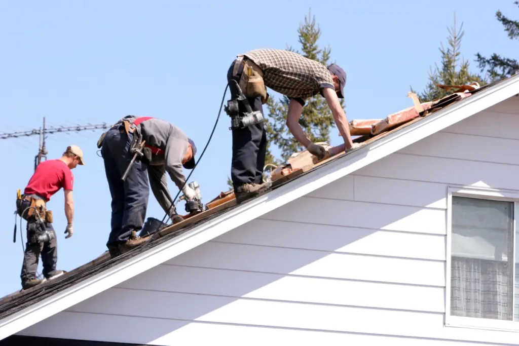 Roofing crew of three workers installing shingles and underlayment along the roofline of a residential house.