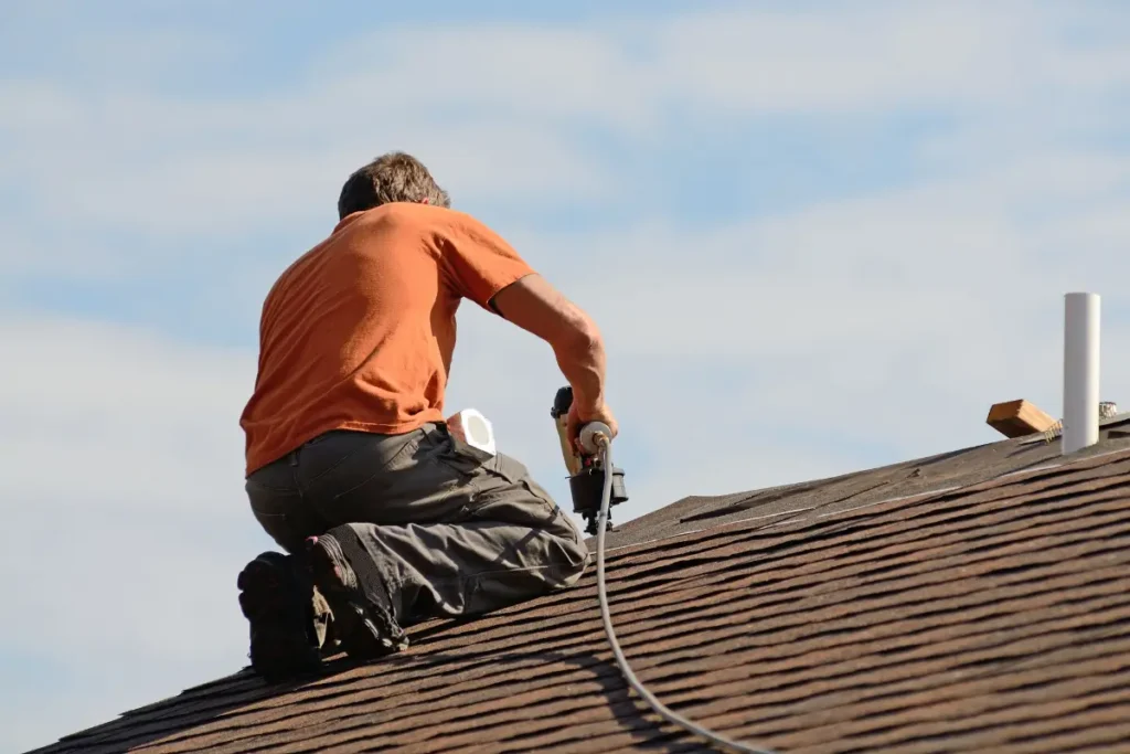 Roofing contractor kneeling on a pitched residential roof using a pneumatic nail gun to install asphalt shingles.