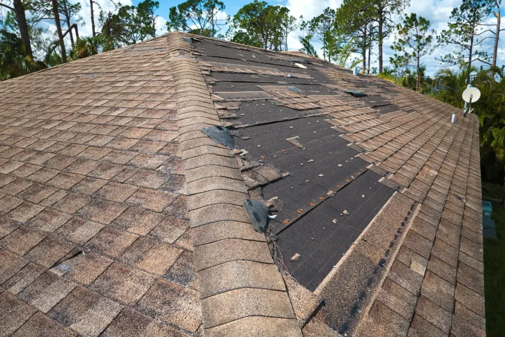 Storm-damaged residential roof showing extensive missing asphalt shingles and exposed black underlayment decking.