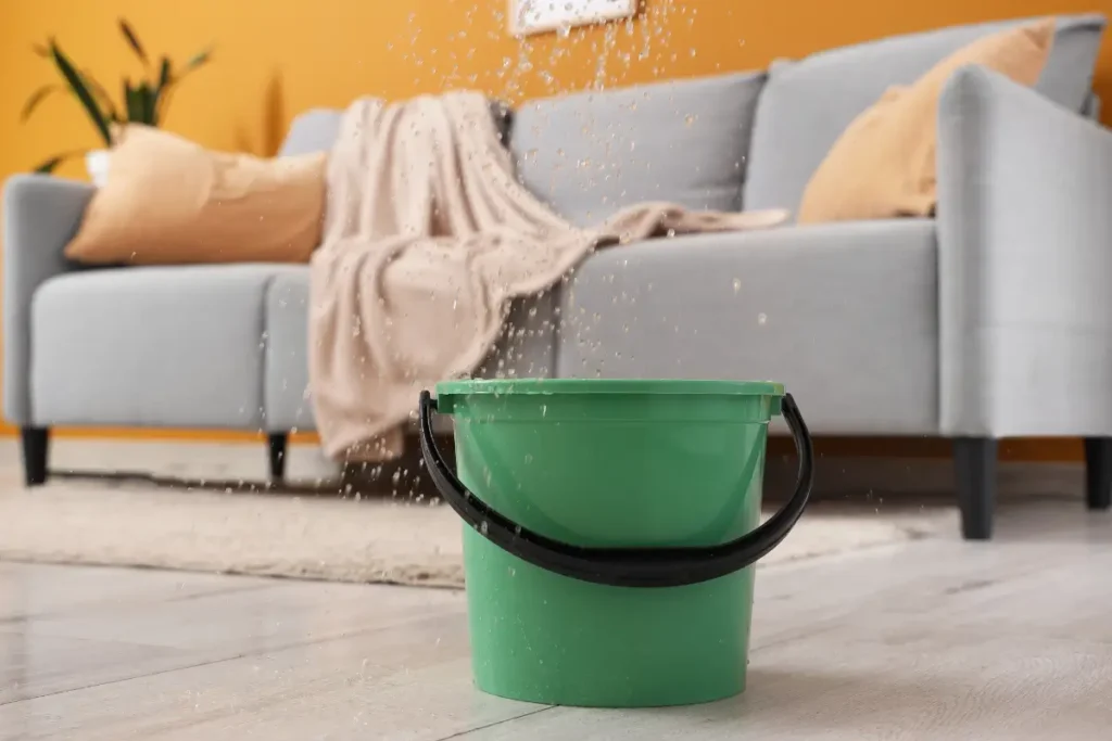 Green plastic bucket catching active ceiling water leaks in front of a grey sofa in a living room.