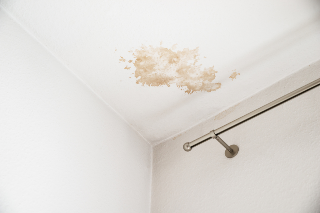 Brown water damage stain on a white ceiling near a curtain rod, indicating a hidden roof leak.