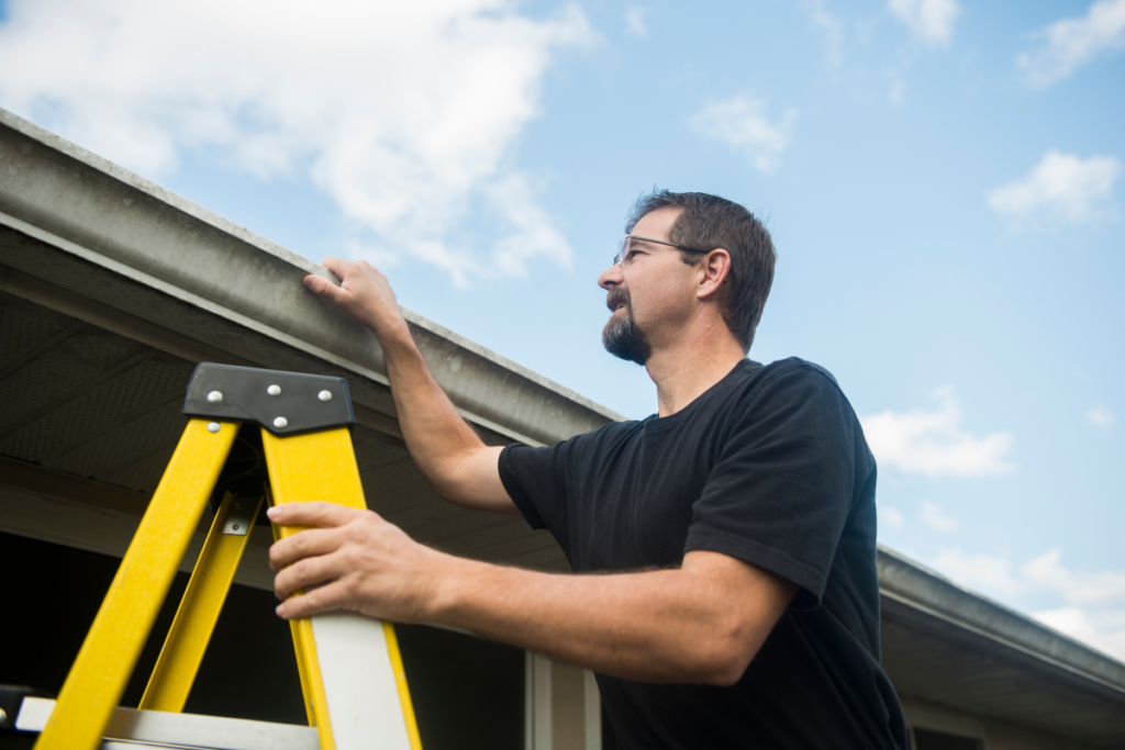 Homeowner standing on a yellow ladder to inspect rain gutters along the roof edge for potential leak causes.