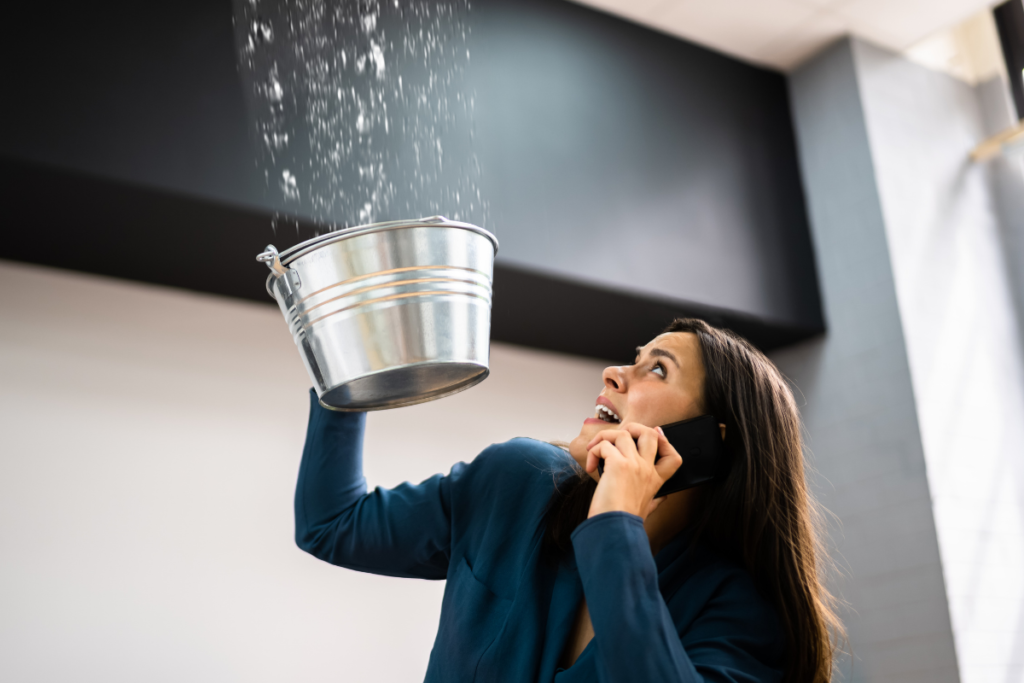 Frustrated woman holding a silver bucket under a severe ceiling leak while calling emergency roof repair services.