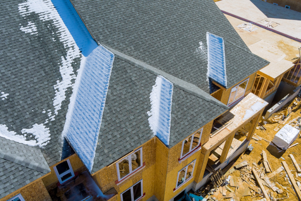 Aerial view of a residential building under construction with grey asphalt shingles, showing snow accumulation in the roof valleys.
